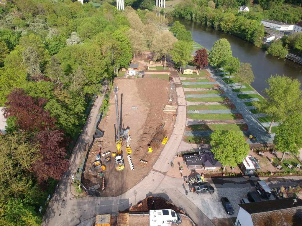 Luftaufnahme einer Baustelle mit Bohrgerät und Baumaterial neben einem Parkplatz am Flussufer, umgeben von Bäumen.