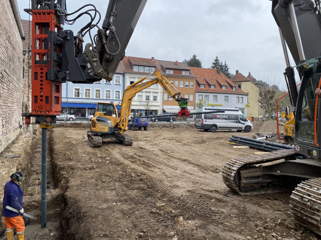 Ein Bagger hält ein Stahlrohr senkrecht neben einer Mauer, während ein zweiter ein weiteres Rohr waagerecht transportiert.